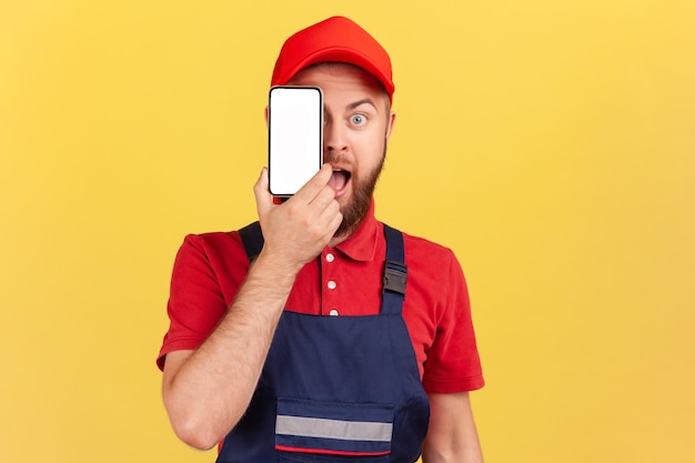 Überraschter Arbeiter in blauer Uniform, der mit einem Handy mit leerem Display steht und das Auge bedeckt