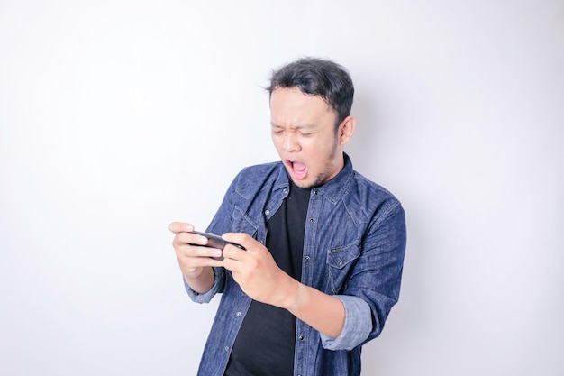 Überraschter asiatischer Mann mit marineblauem Hemd, der auf sein Smartphone zeigt, das durch weißen Hintergrund isoliert ist
