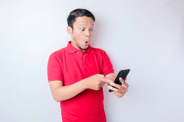 Überraschter asiatischer Mann mit rotem T-Shirt, der auf sein Smartphone zeigt, das durch weißen Hintergrund isoliert ist