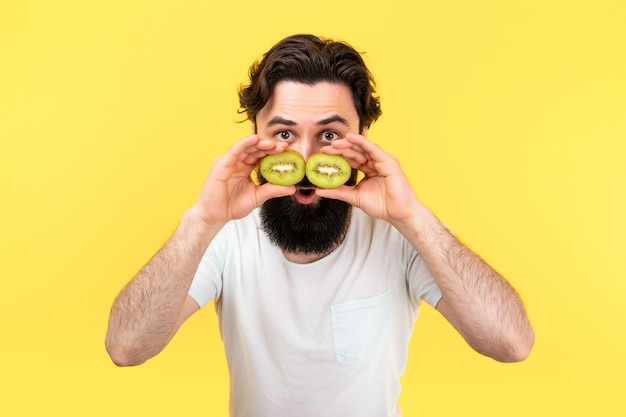 Überraschter bärtiger Mann, der Scheiben grüner Kiwis in Form einer Brille vor die Augen hält