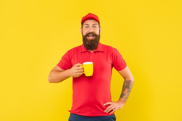 Überraschter bärtiger Mann in lässiger roter Mütze und T-Shirt mit Tasse Tee auf gelbem Hintergrund