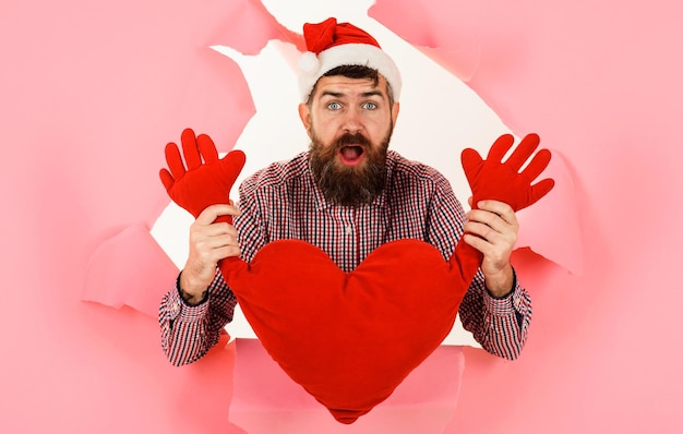 Überraschter bärtiger Mann in Weihnachtsmütze mit Plüschherz Liebe Romantik Valentinstag Geschenkidee Konzept