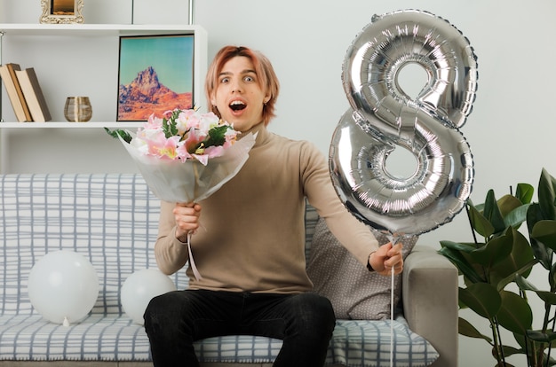 Überraschter hübscher Kerl am glücklichen Frauentag, der Ballon und Blumenstrauß Nummer acht hält und auf dem Sofa im Wohnzimmer sitzt