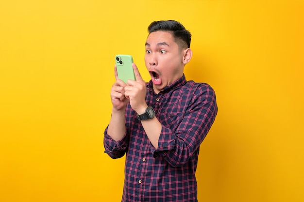 Überraschter junger asiatischer Mann, der auf den Smartphone-Bildschirm schaut und emotional auf Online-Nachrichten reagiert, isoliert vor gelbem Hintergrund