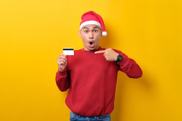 Überraschter junger asiatischer Mann in Weihnachtsmütze, der mit dem Finger auf Kreditkarte auf gelbem Studiohintergrund zeigt Feier Weihnachtsfeiertage und Neujahrskonzept