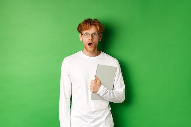 Überraschter junger Mann, der Laptop hält und in die Kamera starrt, Brille und weißes T-Shirt trägt und über grünem Hintergrund steht.