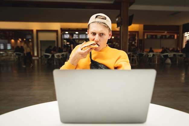 Überraschter junger Mann in Freizeitkleidung und Mütze benutzt Laptop und isst Burger im Café