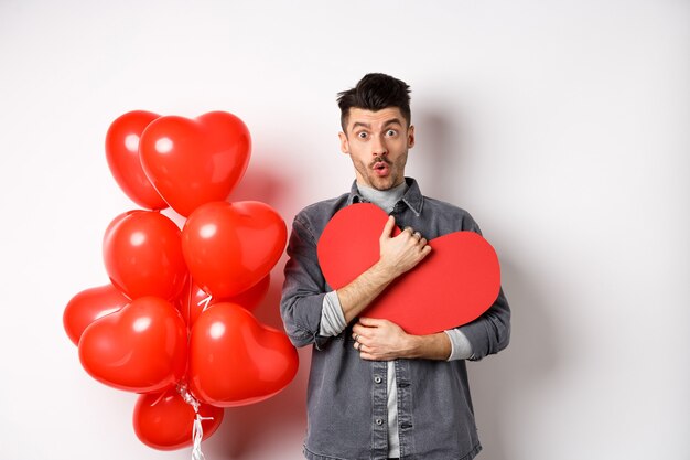 Überraschter Mann, der Valentinsherzkarte hält und wow sagt, erstaunt in die Kamera schaut, erhält geheimes Geständnis am Tag des Liebenden, das nahe romantischen Luftballons auf Weiß steht.