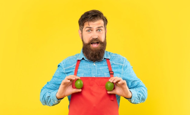 Überraschter Mann in roter Schürze mit frischen Limetten, Zitrusfrüchten, gelber Hintergrund, Gemüsehändler