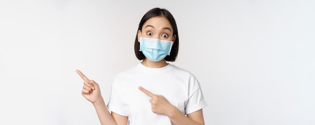 Überraschtes asiatisches Mädchen in medizinischer Maske, das mit dem Finger nach links zeigt und ein Promo-Angebot zeigt, das die Augenbrauen hochzieht, erstaunte Reaktion, die vor weißem Hintergrund steht