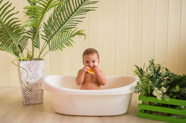 Überraschtes Baby, das im Bad sitzt und eine Orange auf einer Holzwand isst