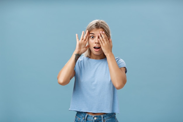 Überraschtes entzücktes Mädchen, das die Hälfte des Gesichts mit der Handfläche bedeckt und mit dem Auge auf Kamera öffnet Mund vor Erstaunen öffnet