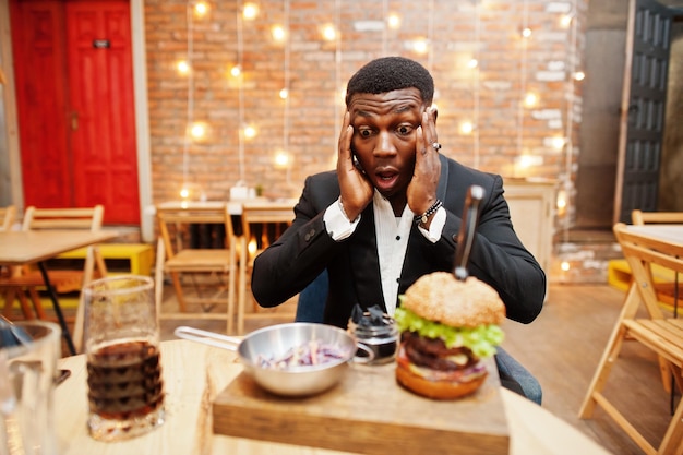 Überraschtes Gesicht eines respektablen jungen Afroamerikaners im schwarzen Anzug, der im Restaurant mit leckerem Doppelburger und Sodagetränk sitzt