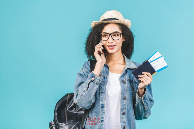 Überraschtes glückliches junges Afroamerikanermädchen lokalisiert auf blauem Hintergrund im Studio. Reisekonzept. Halten Sie Ihren Reisepass und Ihre Bordkarte. Telefon benutzen.