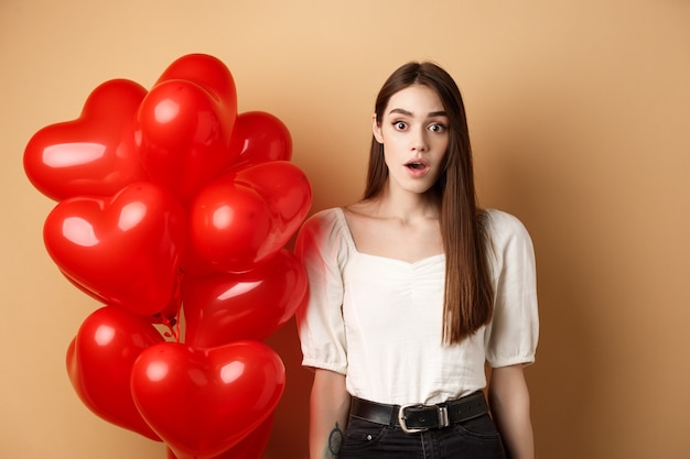 Überraschtes hübsches Mädchen, das nahe Herzballons steht und mit Ehrfurcht in die Kamera schaut.