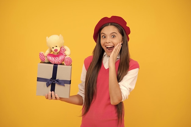 Überraschtes jugendlich Mädchen im französischen Barett halten Spielzeuggeschenk und Geschenkbox auf gelbem Hintergrund Kindheit