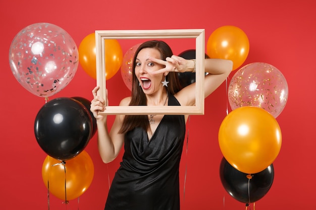 Überraschtes junges Mädchen im schwarzen Kleid, das das Zeigen des Siegeszeichens feiert, das Bilderrahmen auf rotem Hintergrundluftballons hält. Internationaler Frauentag Frohes neues Jahr, Geburtstagsmodell-Feiertagsparty-Konzept.