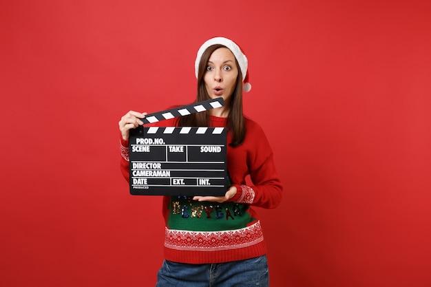 Überraschtes junges Santa-Mädchen, das den Mund weit offen hält und die klassische schwarze Filmklappe hält, die auf rotem Hintergrund isoliert ist. Frohes neues Jahr 2019 Feier Urlaub Party Konzept. Kopieren Sie Platz.