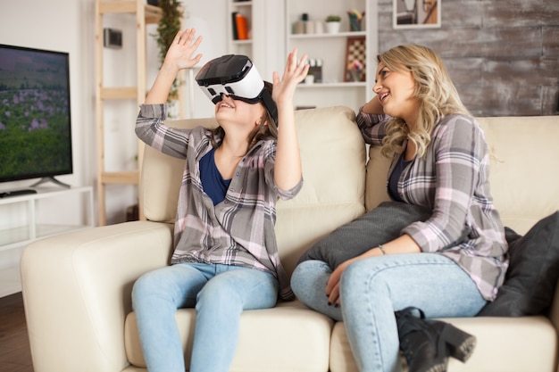 Überraschtes kleines Mädchen, das neben ihrer Mutter auf der Couch sitzt, während sie ein Virtual-Reality-Headset verwendet.