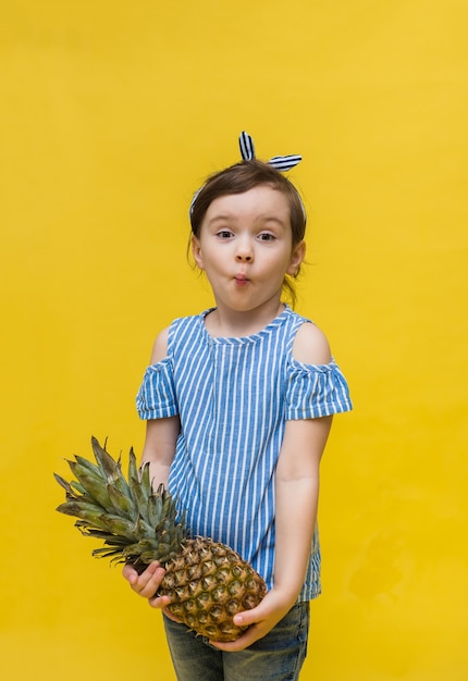 Überraschtes kleines Mädchen in einem Stirnband und einem gestreiften T-Shirt hält eine Ananas an einer gelben Wand mit Platz für Text