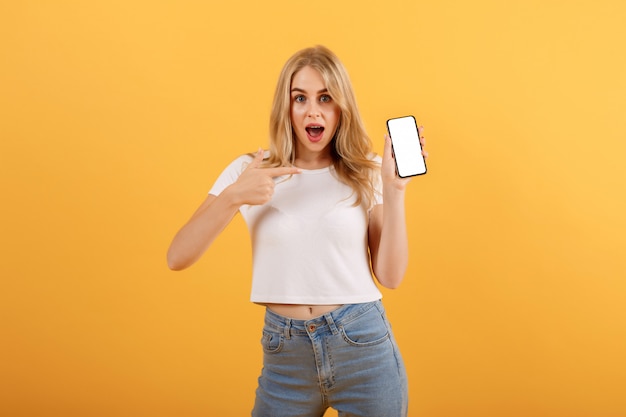 Überraschtes Mädchen im weißen T-Shirt ist auf einem orangefarbenen Hintergrund und zeigt ihren Finger auf den Smartphone in ihrer Hand