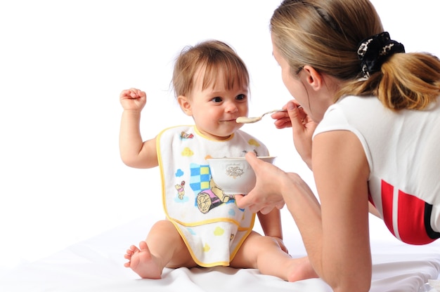 Überraschtes süßes kleines Baby, das Essen vom Löffel isst. Mutter, die Platte hält und Kind füttert. Isoliert auf weißem Hintergrund