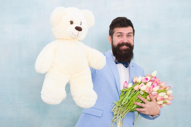 Überraschung für sie Mann bärtiger Gentleman Anzug Fliege halten Teddybär und Blumenstrauß Gentleman macht romantische Überraschung für sie Gentleman romantisches Geschenk Geburtstagsgrüße Alles Gute zum Geburtstag, meine Liebe