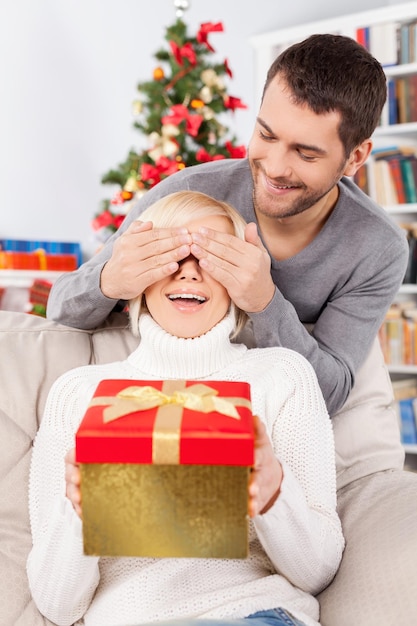 Überraschung! Schöne junge Frau, die auf der Couch sitzt und eine Geschenkbox hält, während ihr Freund hinter ihr steht und ihre Augen mit den Händen bedeckt