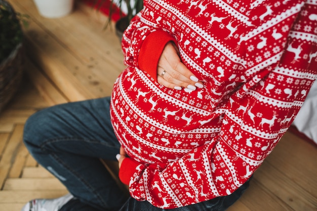Übersteigen Sie hinunter Bauch einer schwangeren Frau, die rote dekorative Weihnachtsstrickjacke trägt