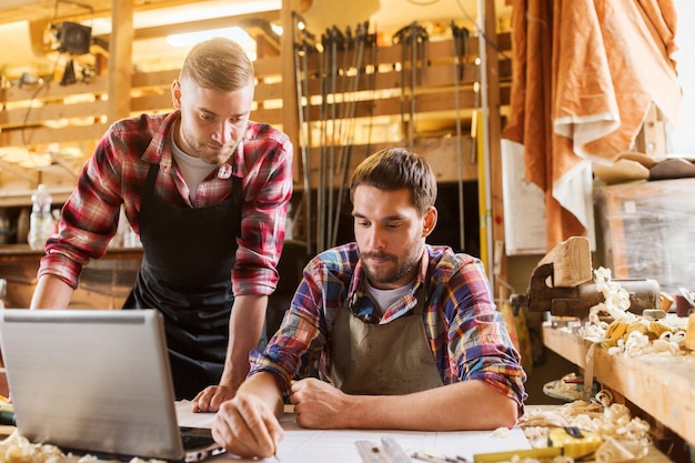 Foto beruf, zimmerarbeit, technologie und menschenkonzept - zwei zimmerleute mit laptop-computer und blaupause in der werkstatt