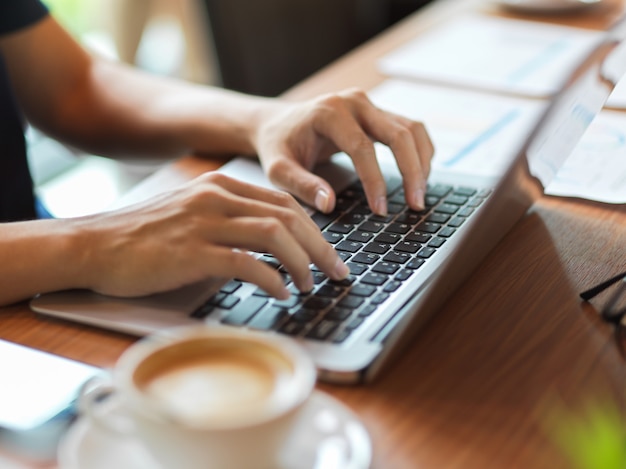 Berufstätige Frau, die auf der Laptop-Tastatur mit Finanzbericht und Kaffeetasse daneben auf dem Schreibtisch tippt