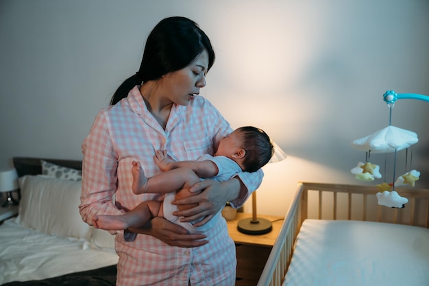 Überwältigte asiatische neue Mutter, die neben dem Babybett steht, streichelt ihr weinendes neugeborenes Mädchen, während sie versucht, sie um Mitternacht im Schlafzimmer zu beruhigen.