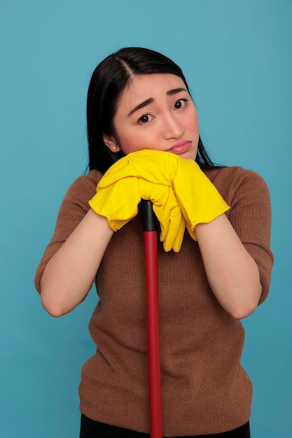 Überwältigte traurige asiatische Hausfrau, die am Bürstenstab gelbe Handschuhe hält, die am Bürstenstab festhalten, Hausreinigungskonzept, Haushälterin, die sich negativ erschöpft fühlt und sich etwas ausruht