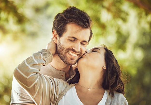 Besar el amor y la pareja en el parque sonríen y se divierten juntos al aire libre Día de San Valentín romance y cuidado afecto e intimidad de un hombre y una mujer felices besándose en la mejilla en una cita romántica al aire libre