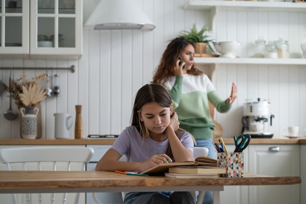 Beschäftigte Mutter, die kleine Tochter ignoriert, genießt Handygespräche, wenn trauriges Schulmädchen Hausaufgaben lernt