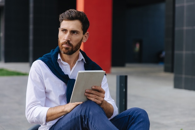 Beschäftigter bärtiger Mann in stilvollem Outfit, der vor seinem Büro an einem digitalen Tablet arbeitet. Junger Geschäftsmann, der auf Bank sitzt und modernes Gerät verwendet.