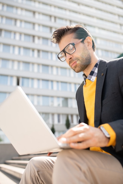 Beschäftigter junger eleganter Unternehmer mit Laptop, der durch Websites im Netz schaut, während er in der städtischen Umgebung arbeitet