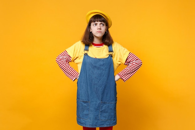 Beschäftigter Mädchen-Teenager in französischer Baskenmütze, Denim-Sommerkleid, das mit in die Hüfte gestemmten Armen steht, isoliert auf gelbem Hintergrund im Studio. Menschen aufrichtige Emotionen, Lifestyle-Konzept. Mock-up-Kopienbereich.