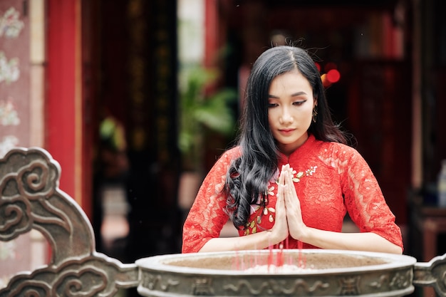 Bescheidene junge hübsche VIetnamesin, die im buddhistischen Tempel betet, während sie neben einer bronzenen Urne mit brennenden Räucherstäbchen bleibt