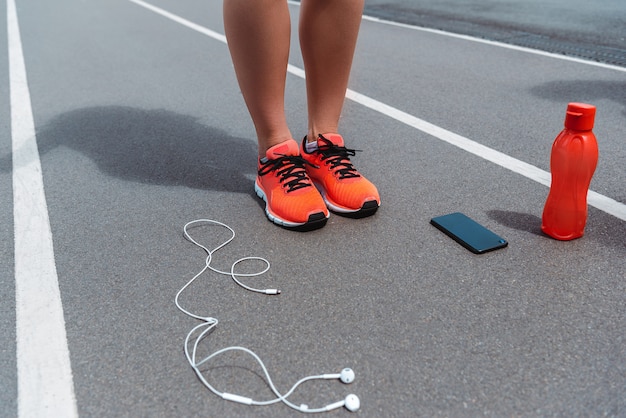 Beschnittene Ansicht der weiblichen Beine in den Turnschuhen auf Laufbahn nahe Smartphone und Kopfhörer