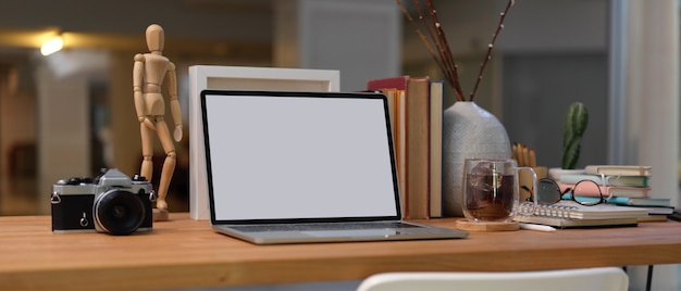Beschnittene Aufnahme des Arbeitsbereichs mit Laptop mit leerem Bildschirm, Büchern, Verbrauchsmaterialien und Dekorationen auf Holzschreibtisch im Wohnzimmer