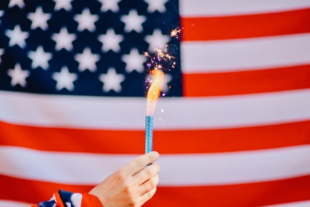 Beschnittene Hand hält angezündetes Feuerwerk gegen die amerikanische Flagge