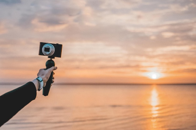 Beschnittene Hand hält eine Kamera am Strand gegen den Himmel während des Sonnenuntergangs