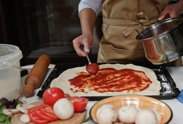 Beschnittene italienische Hausfrau in beiger Kochschürze, die frisch zubereitete Tomatensauce auf ausgerolltem Teig in Pizzapfanne verteilt