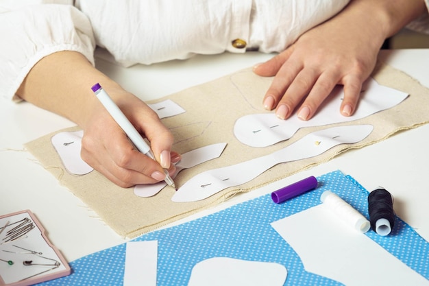 Beschnittene weibliche Näherin zeichnet Konturen mit einem Stift aus festgestecktem Papier Nähmuster auf Textilfolie auf dem Tisch Spielzeugkleidung