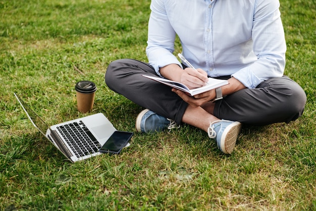 Beschnittener kaukasischer Mann in Geschäftskleidung, sitzend auf Gras im Park mit gekreuzten Beinen und Aufschreiben von Notizen im Notizbuch während der Arbeit am Laptop