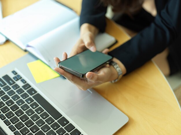 Beschnittener Schuss der Geschäftsfrau, die Smartphone auf Arbeitsbereich mit Laptop und Zeitplanbuch im Büroraum verwendet