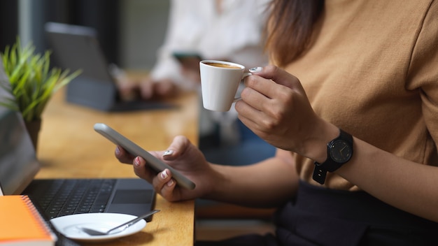 Beschnittener Schuss der jungen Frau unter Verwendung des Smartphones beim Halten einer Tasse Kaffee