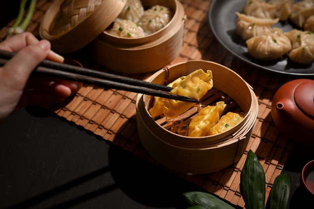Beschnittener Schuss der weiblichen Hand mit Essstäbchen, das Dimsum Knödel im Bambusdampfer auf Esstisch pflückt