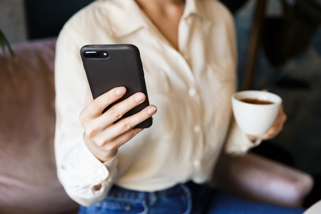 Beschnittenes Bild der hübschen schönen jungen Frau, die im Café drinnen arbeitet, trinkt Kaffee mit Handy.